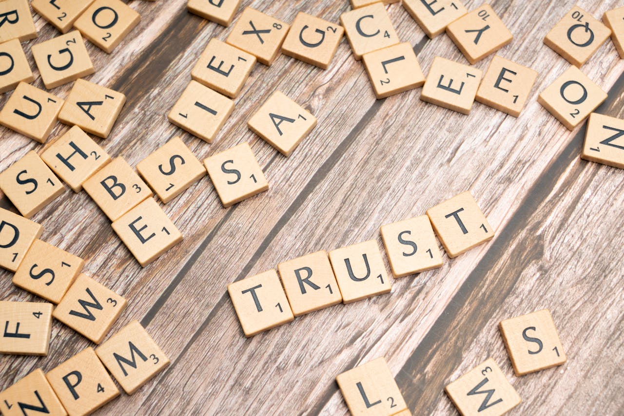 Scrabble letter tiles form the word 'Trust' on a wooden table, surrounded by scattered tiles.